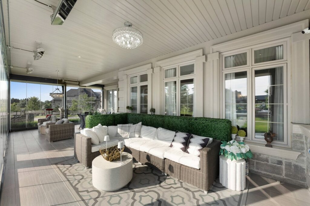 A covered patio with a wicker sofa, white cushions, round coffee table, potted plants, and large windows overlooking a landscaped yard.