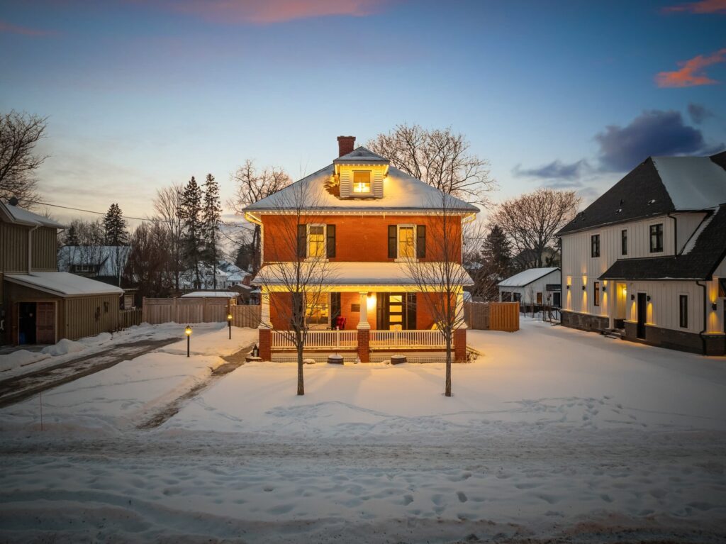A two-story brick house with a front porch is illuminated at dusk, surrounded by snow and flanked by neighboring homes and leafless trees.