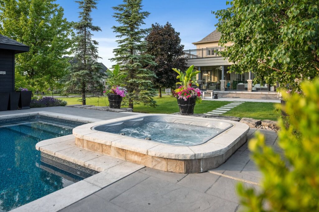A backyard with a hot tub next to a swimming pool, surrounded by stone patio, plants, and outdoor seating near a house.