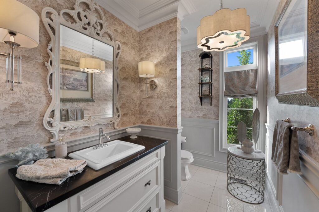 Elegant bathroom with ornate mirror, patterned wallpaper, single sink vanity, wall sconces, round table, towels, and a window letting in natural light.