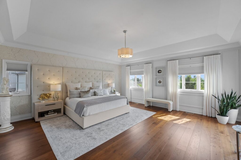 Spacious bedroom with a large bed, two nightstands, a white bench, hardwood floors, large windows with white curtains, and potted plants.