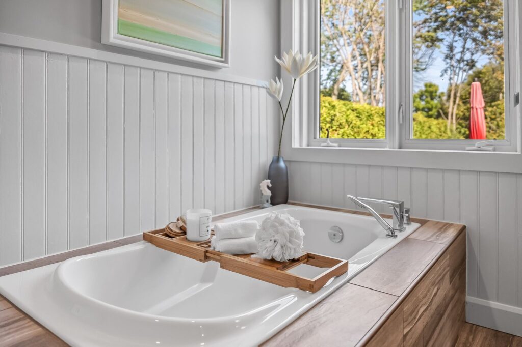 A bathtub with a wooden tray holding a towel, soap, and a mug is situated under a window with white walls and a vase of white flowers.