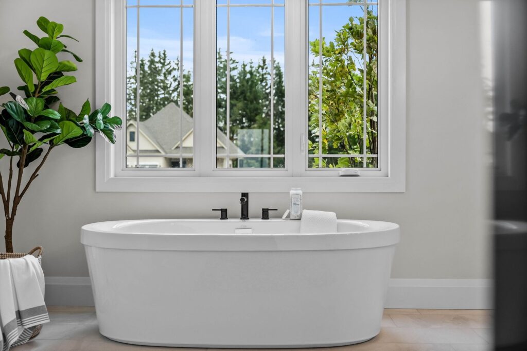 A white freestanding bathtub sits below large windows with a view of trees and a house. A towel and plant are nearby, giving the bathroom a bright and clean look.
