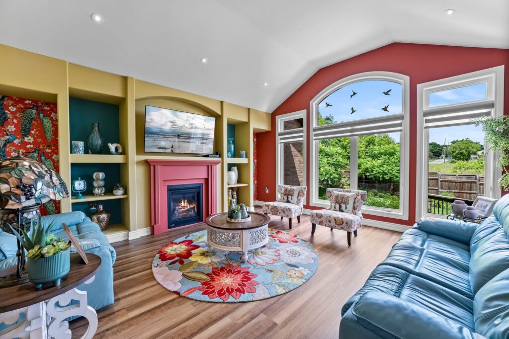 A colorful living room features a blue sofa, round floral rug, fireplace with pink mantel, large arched window, built-in shelves, and decorative accents.