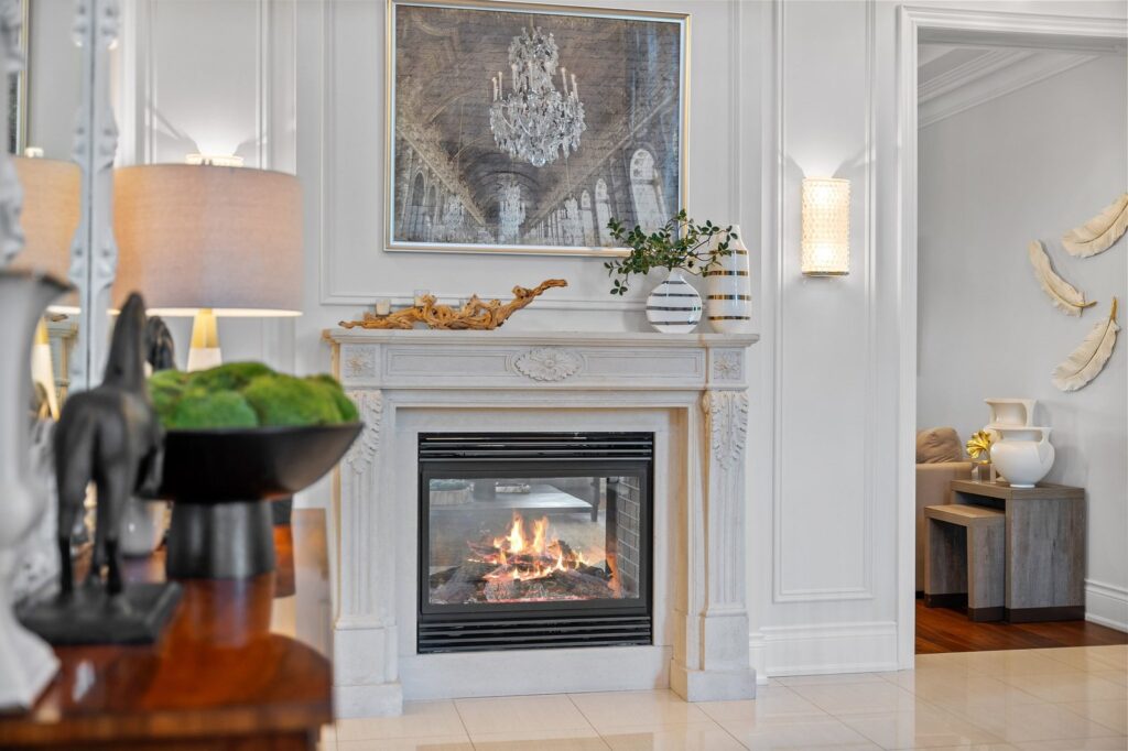 A lit fireplace with a stone mantel is centered below a framed artwork. Decorative items and a plant are on the mantel; a table with ornaments is in the foreground.