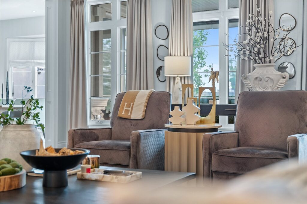 A living room with two gray armchairs, a round side table, large windows with beige curtains, and decorative vases and ornaments on the table and windowsill.