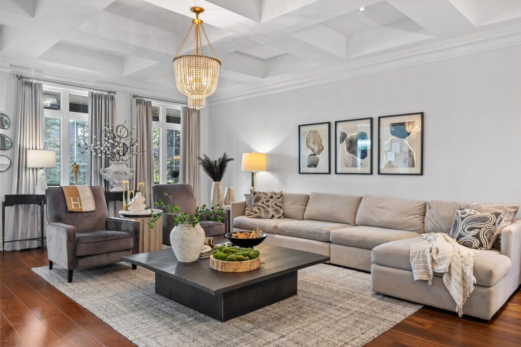 A modern living room with a beige sectional sofa, two gray armchairs, a large black coffee table, wall art, and a chandelier on a coffered ceiling.