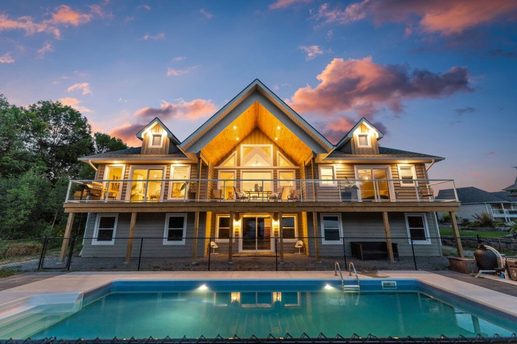 Modern two-story house with large windows and balcony, illuminated at dusk, with a swimming pool in the foreground and trees in the background.