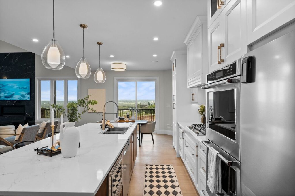 Modern kitchen with white cabinets, marble island, pendant lights, built-in ovens, and large windows overlooking an outdoor view.