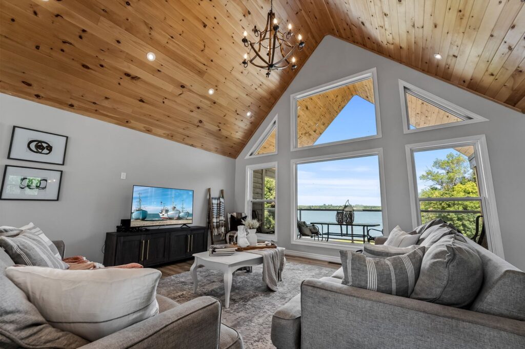 A living room with vaulted wood ceiling, large triangular windows overlooking a lake, neutral-toned furniture, a TV, and a chandelier.
