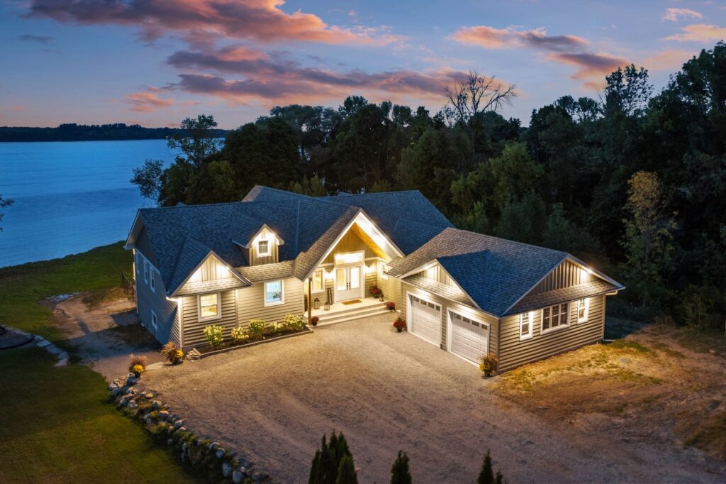 A modern, single-story house with exterior lights on sits beside a large lake, surrounded by trees, during sunset.