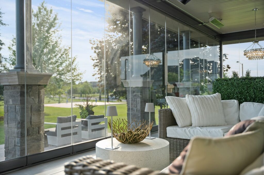A modern patio with glass walls, wicker furniture with cream cushions, a round table, and outdoor chandeliers overlooking a landscaped lawn.