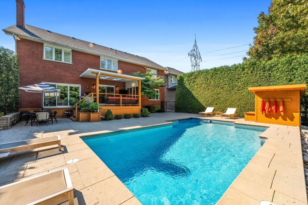 A backyard swimming pool with lounge chairs, a shaded sitting area, and a red brick house in the background under clear blue sky.