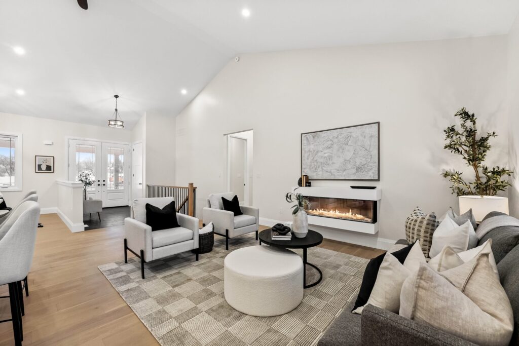 Modern living room with light walls, armchairs, a gray sofa, round ottoman, wall-mounted fireplace, and large area rug. Artwork and a potted plant decorate the space.