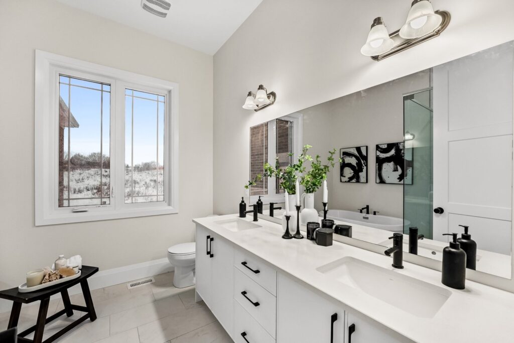 Modern bathroom with a double sink vanity, black fixtures, wall-mounted lights, a large mirror, a window, and minimalist decor including green plants and monochrome art.
