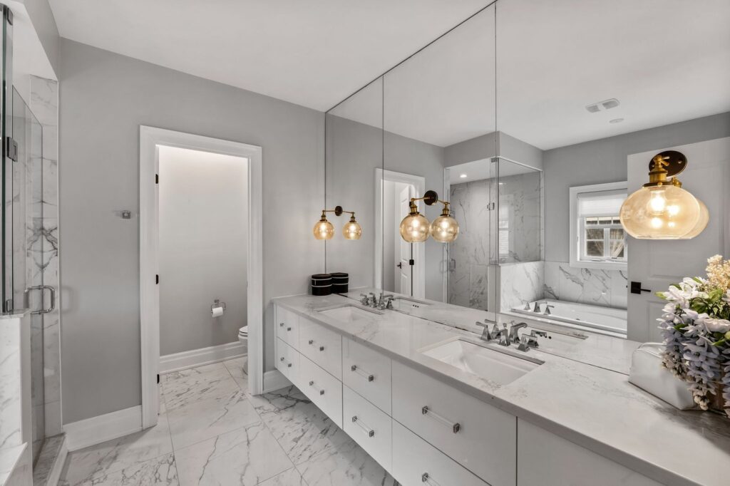 Modern bathroom with double sinks, large mirror, marble floor and walls, two pendant lights, and a glass shower enclosure. A toilet room is visible through an open door.