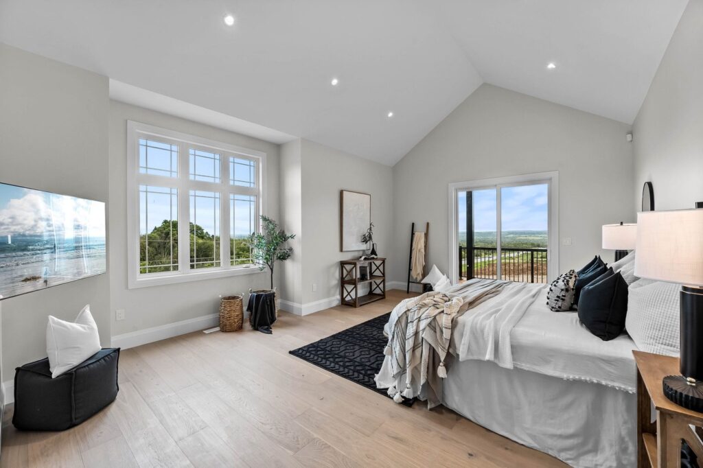 Modern bedroom with large windows, a bed with white linens, a black rug, desk, decorative plants, and a view of the outdoors through sliding glass doors.