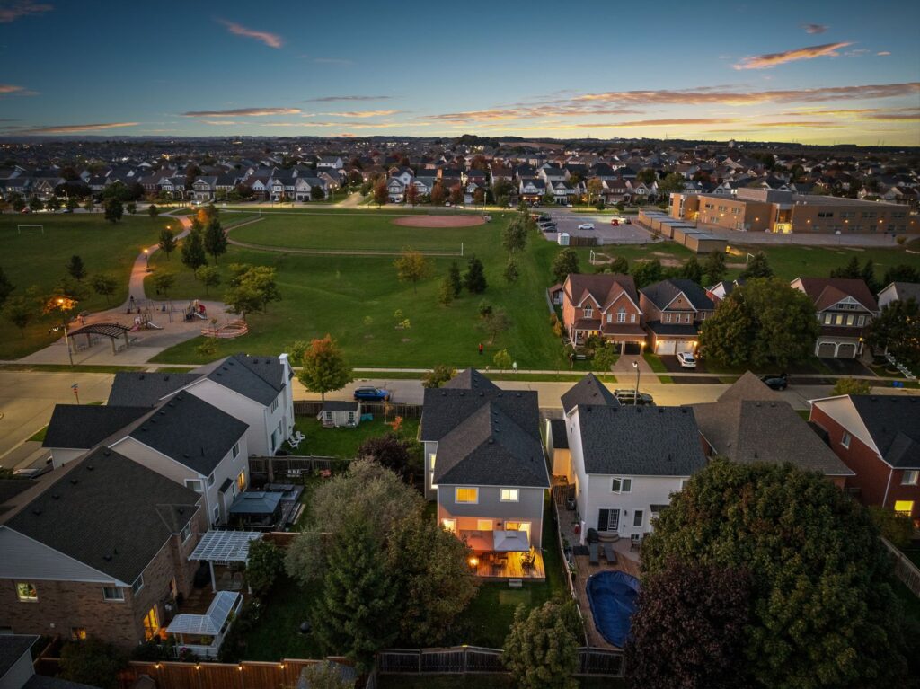 Aerial view of a suburban neighborhood at dusk showing houses, a park with a playground, a baseball field, and nearby buildings under a partly cloudy sky.
