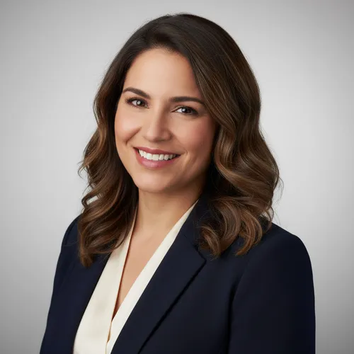 A woman with medium-length brown hair wearing a dark blazer and light blouse, smiling at the camera against a plain light gray background.