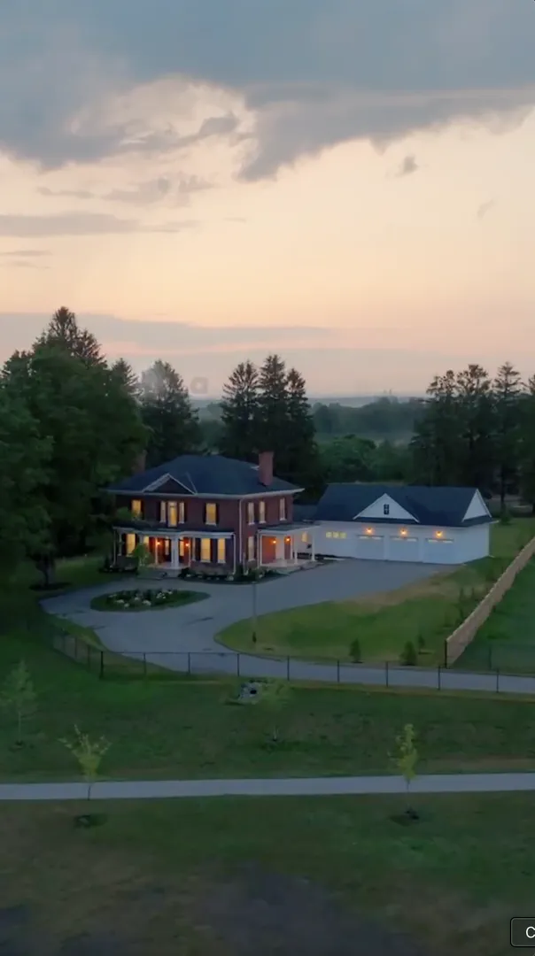 Two-story brick house with white columns and a large driveway, adjacent to a detached garage, surrounded by trees and a fenced yard at dusk.