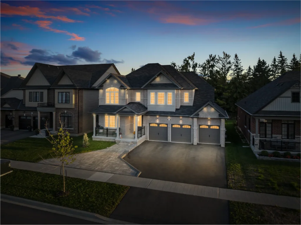 A two-story suburban house with lit windows, a three-car garage, and a driveway at dusk, situated between two similar homes.