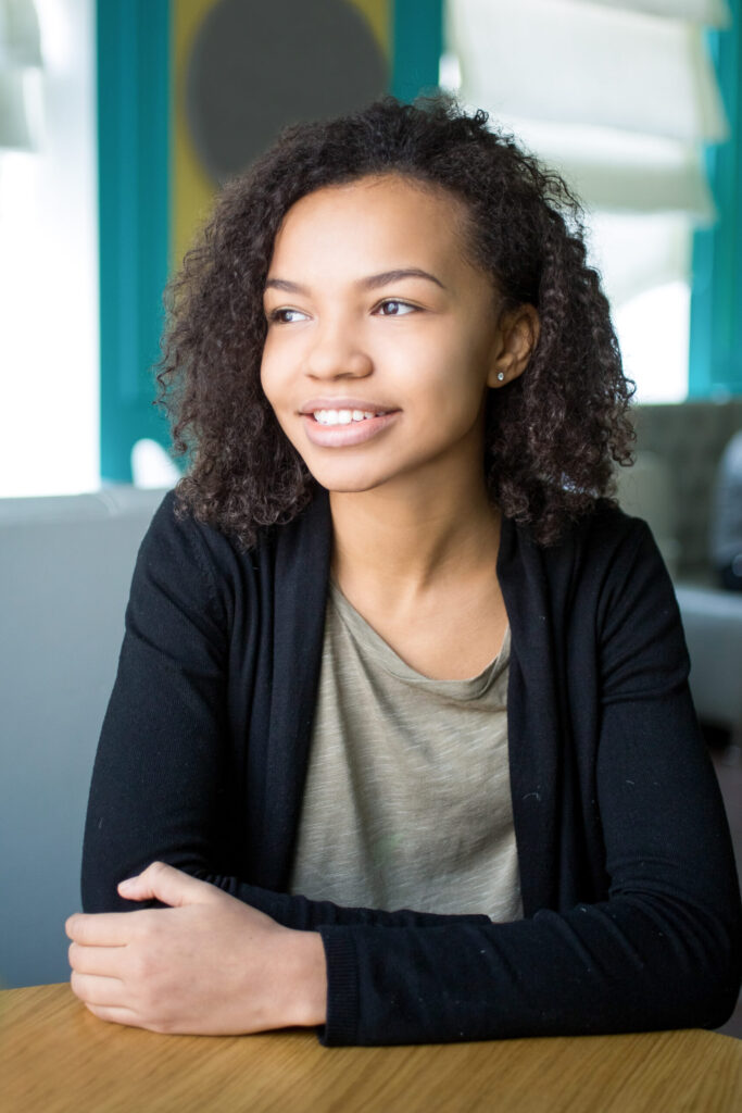 A young woman with curly hair sits at a table, looking to the side and smiling. She is wearing a green shirt and a black cardigan.
